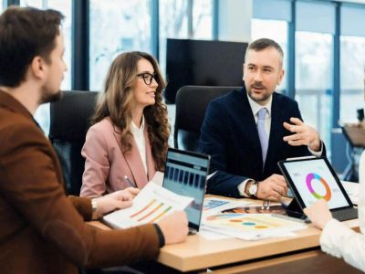 Four business professionals discuss charts and graphs during a meeting at a conference table with laptops and documents.