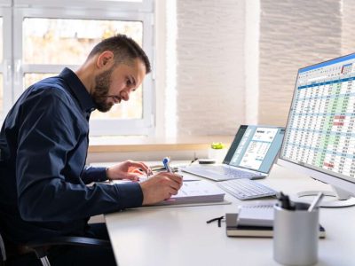 Man working at a desk with financial spreadsheets on a computer monitor, laptop, and documents in a bright office.