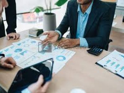 Three people in business attire discussing charts and graphs at a desk with documents and a tablet.